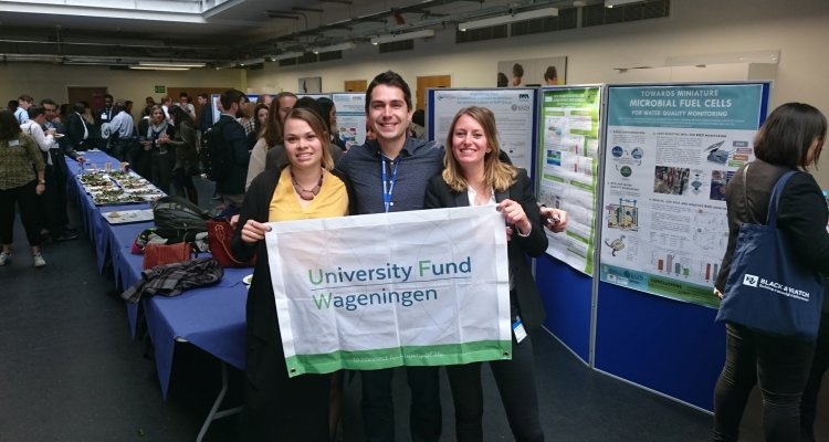 Three students at a conference with a flag with the logo of University Fund Wageningen on it