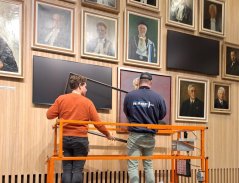 Two workmen standing on a hydraulic lift looking at the portrait paintings from close by.